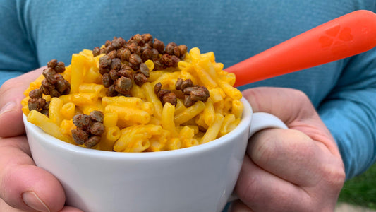 A close-up photograph of two male hands holding a cup brimming with mac n cheese topped with crunchy toasted lentils