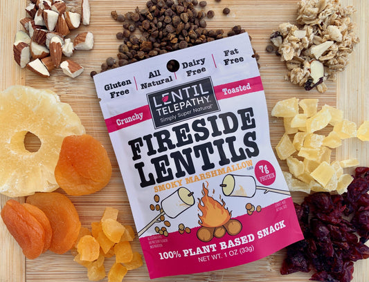 A package of Lentil Telepathy crunchy toasted lentil snacks in smoky marshmallow flavor arranged on a wooden snack board alongside almond pieces, dried fruit, and granola