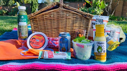 A large wicker picnic basket set atop a colorful woven blanket, surrounded by snacks: Lentil Telepathy, Suja Juice, Bitchin Dip, Perfect Bar in Mint Chocolate, Belching Beaver IPA can, and Chosen salad dressing next to a small salad